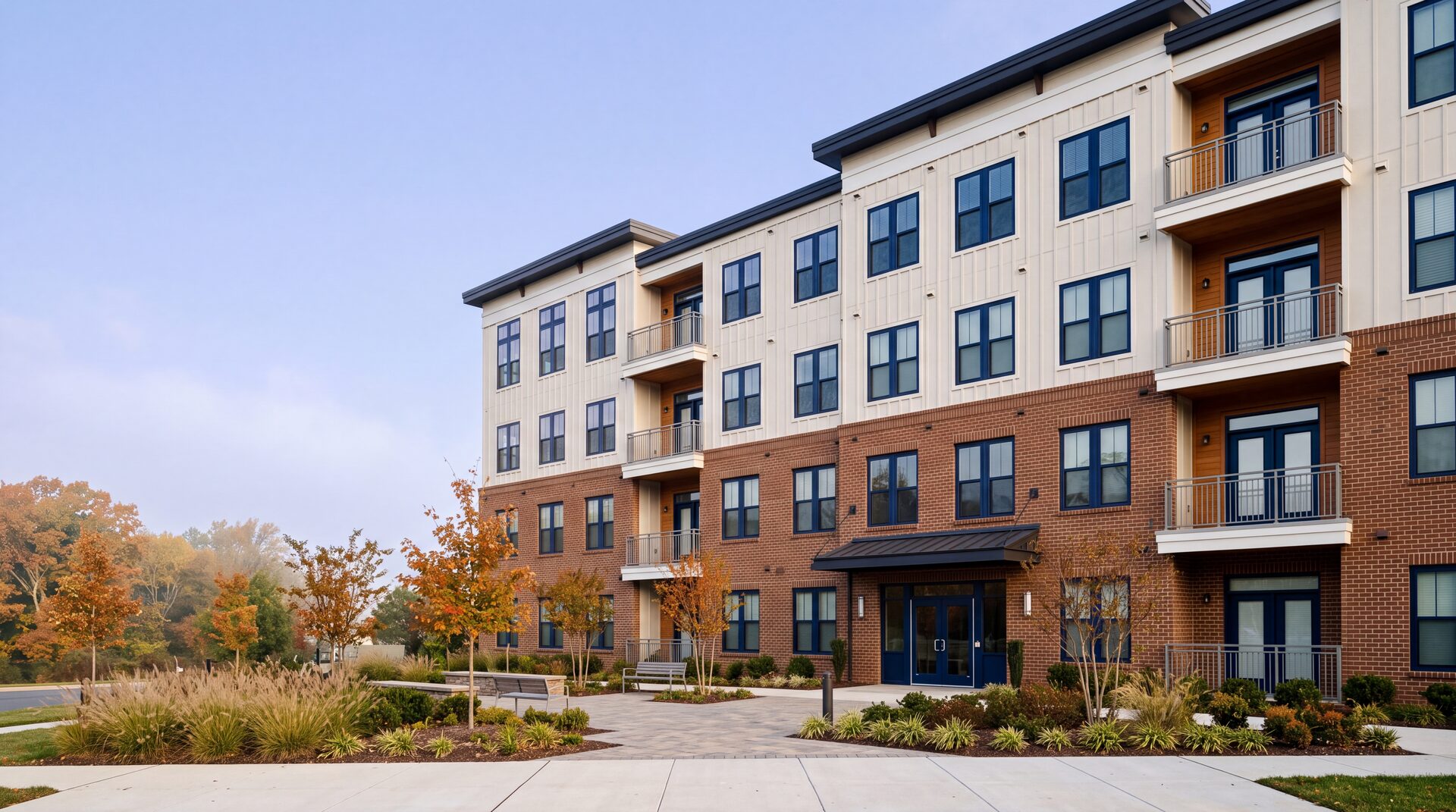 A newly completed four-story brick and white-sided apartment building with balconies and a landscaped entry plaza in autumn light